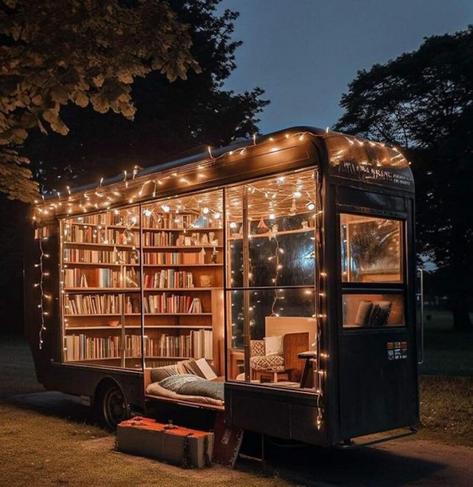A pull along trailer that used to be enclosed, now has windows all over and inside is a library of books lining the walls. On the floor is a mat and setup to sleep, and in the corner a comfy chair.  Little string lights line the top and sides of most of the windows while a large block of wood stands at the side as a stair.