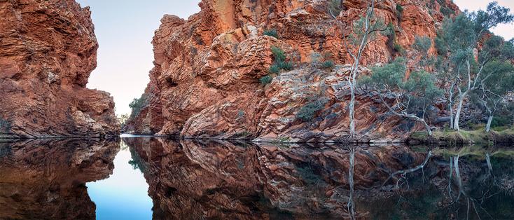 Folded rock layers and river gums are reflected in the calm surface of an outback waterhole.