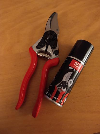 Eine Rebschere bzw. Rosenschere mit roten Griffen liegt zusammen mit einer Sprühdose Pflegespray auf einem hellen Holztisch.