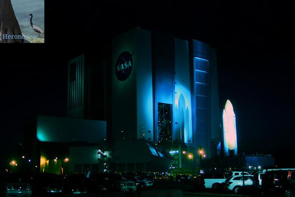 An outdoor, nighttime photograph of the Space Shuttle stack, with the orange External Tank and white Solid Rocket Boosters facing the camera, hiding the plane-like Shuttle. Bright light focus on the External Tank and cast shadows on the large, gray building and the tall doors behind the Shuttle.  There are cars and trucks parked closer to the camera with the Shuttle in distance.