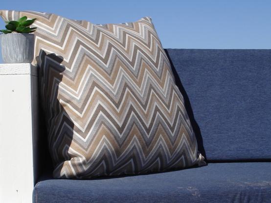 Beach sofa with navy blue seating cushions. A decorative pillow with zigzag pattern sits in the corner. On top of the armrest sits an artificial plant.