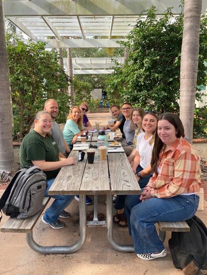 Group of scientists sitting around a picnic table outdoors, drinking coffee and scheming about experiments