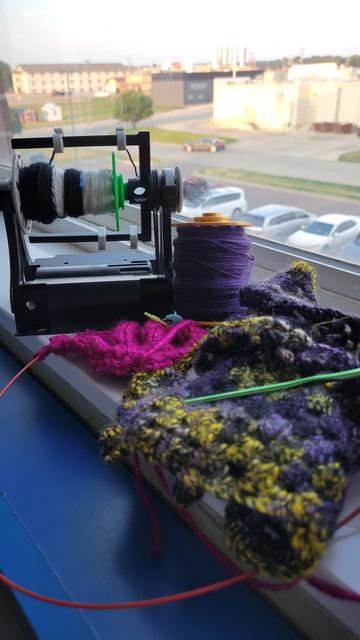 Parking lot and buildings out the window in La Mars Iowa, with an Electric Eel Wheel Nano 2 spinner, purple bobbin of singles, pink knitted sweater, and enby colored crochet project.