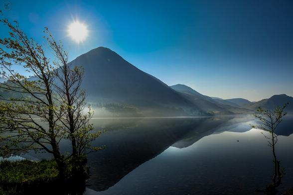 Shot across very still lake waters, with trees on left frame and mountains in background, clearly reflected in the water. Sun and blue skies above