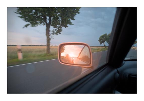 Weitwinkelaufnahme eines Außenspiegels am Auto durch die Scheibe. Im Rückspiegel ist der Sonnenuntergang zu sehen. Um den Rückspiegel herum eine durch die Bewegung unscharf abgebildete Landstraße und Felder.