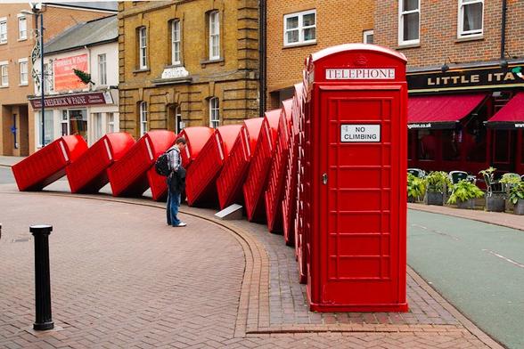 An image captured in the Royal Borough of Kingston upon Thames, showcasing a line of twelve red phone booths. The phone booths are arranged in a domino-like manner, leaning towards each other as if about to topple over