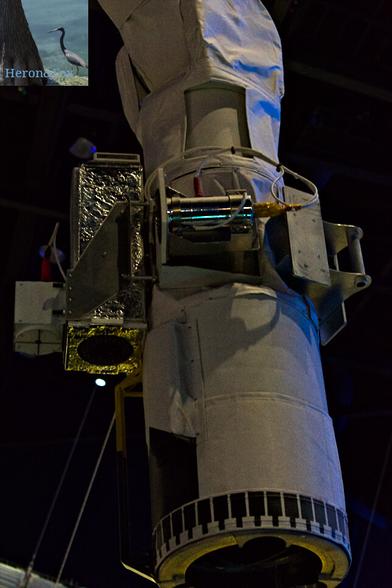 An indoor photograph of a vertical white cylinder with an opening at its bottom and with cameras and electronics near the top. The top attaches to a mechanical joint covered in white fabric-like material.