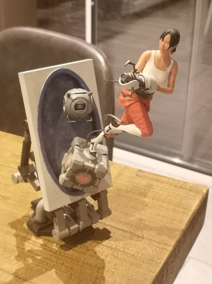 photo d'une statuette miniature (environ 15cm de haut) imprimée en 3D du personnage de Chell, de la série de jeux vidéos Portal, jaillissant d'un portail juchée sur un Cube de Voyage, portant en main le générateur de portails. La sphère Whesley la suit. Le tout a été imprimé et peint par mes petites mimines.