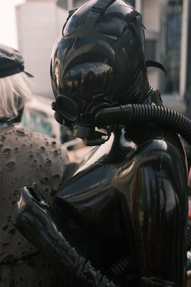 ToyDolly as a figure in a black latex catsuit looks back towards the camera. They are wearing an MSA Millenium gas mask and there is an air hose going to a bubbler bottle which is out of shot.