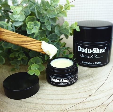 Ein Foto vom einem kleinen und einem großen Glasbehälter mit Sheabutter von Dudu-Shea. Auf einem Spatel aus Olivenholz ist etwas feste Sheabutter an der Spitze zu sehen.