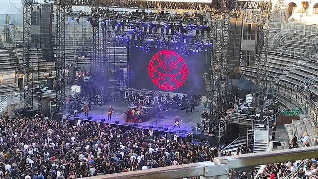 Photo de la scène des arènes de Nîmes pendant le concert d'Avatar. On y voit un bout de la foule en fosse, les membres du groupe et leurs installations lumineuses.