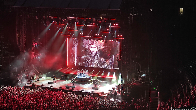 Photo de la scène des arènes de Nîmes pendant le concert de Gojira. On y voit un bout de la foule en fosse, les membres du groupe et leurs installations lumineuses. En arrière plan il y a une image d'une femme Amazone.