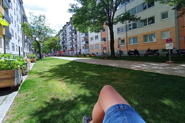 Blick vom Holzstuhl, auf dem ich sitze, über meine Haxn hinweg in die Sommerstraße. Statt Parkplätzen und Autoverkehr sieht man einen Kiesweg, Rasen und Pflanzenkübel.