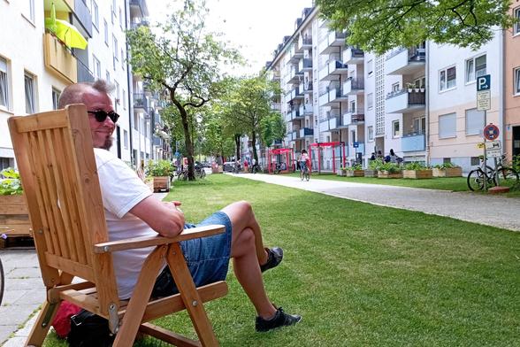 Moi mit Sonnenbrille, weißem T-Shirt, über dem Knie endenden Jeansrock, dünner hautfarbener Strumpfhose und blauen Sneakers auf Holzstuhl mit Blick auf Sommerstraße Kolumbusstraße mit Wiese und Kiesweg.