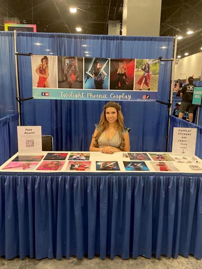 A woman cosplaying as Margaery Tyrell from Game of Thrones sits at a convention booth. The banner behind her says twilight Phoenix Cosplay.