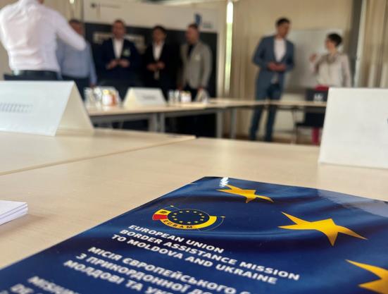 Blick in die Unschärfe in einem Besprechungsraum. Vorne auf dem Tisch liegt ein Papier der EU Border Assistance Mission to Moldova and Ukraine, weiter hinten unterhalten sich fünf Personen.