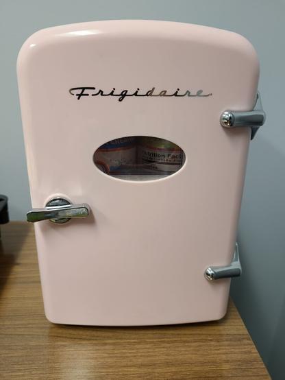 Photograph of a small, 4 can, pink desktop fridge. It is Fridgidare brand, has a silver handle, and a small window where you can see the front two cans.