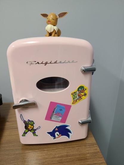 Photograph of a small, 4 can, pink desktop fridge. It is Frigidaire brand with a silver handle and a small window where you can see the first two cans. There are stickers on it from Aespa, a Hammer Brother from Mario, Sonic the Hedgehog, and Leonardo from the Ninja Turtles.