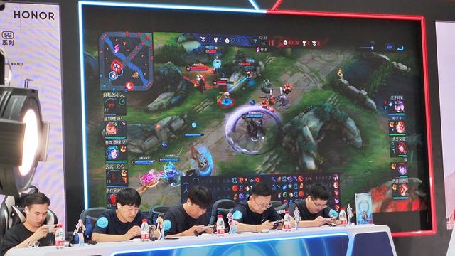 A team of Chinese gamers sit in front of a large monitor displaying an e-sports match of the Chinese mobile MOBA Honor of Kings. On the screen a skirmish begins in the middle lane. On the left side of the screen is displayed an ad for the tournament sponsor, the phone brand Honor.