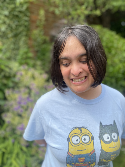 Close up of Raana, a woman with brown skin and black hair cut in a Bob style, in the garden. She is gazing down and smiling. She wears a blue t shirt with two Minions on the front. Behind her are green plants.