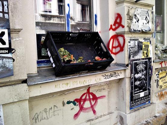 Auf dem blechenen Fensterbrett eines mit Grafitti und Plakaten dekorierten Hauses stehtm schräg an die Fenster gelehnt, eine schwarze Plastekiste. In der Kiste sind Plattpfirsiche, Möhren und Salat erkennbar. Am Fenstersims ist "ALL COPS ARE INCEST ACAI" und Hammer und Sichel mit roter Farbe sowie zwei umkreiste A's erkennbar.
In der Fensterscheibe hinter der schwarzen Kiste spiegelt sich das Haus, das auf der gegenüberliegenden Straßenseite steht.