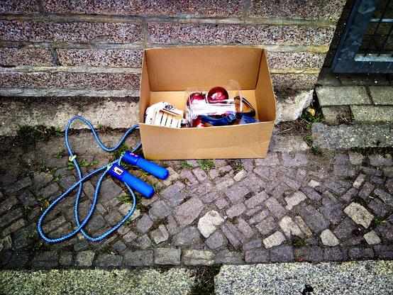 Vor einer Mauer steht auf einem Kleinkopfpflaster-Bürgersteig eine Kiste aus brauner Pappe. Vor der Kiste liegt zusammengeringelt ein blaues Springseil mit einem, in einen der beiden Griffe integrierten, Zähler. In der Kiste sind rote Weihnachtskugeln und ein kleines Holzkistchen erkennbar. Außerdem der Oberteil einer bläulichen Sandale.