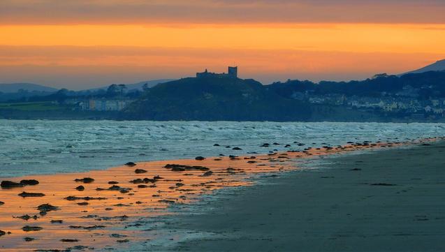 A cloud-streaked orange sky above the shadowy skyline of Criccieth, dominated by its castle. The colour of the sky is reflected in the wet sand of the beach in the foreground.