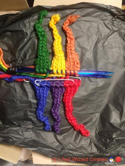 Picture of six knitted cones, red/organge/yellow/green/blue/purple, lying flat on a black tissue paper background. text in the lower left corner reads: <|:-) Red Wizard Creates 🧙‍♂️🔴