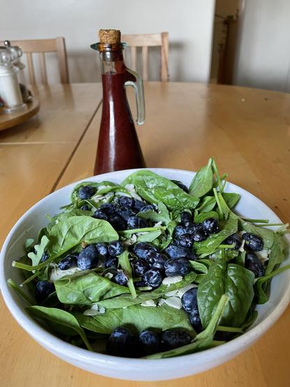 A green salad in a white bowl topped with dark, purple berries. A glass jar of deep maroon salad dressing is in the background.