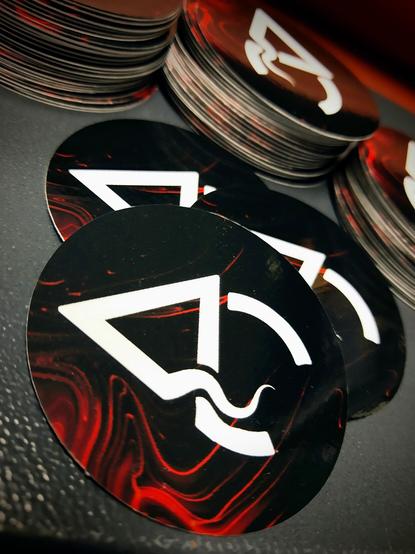 three stacks of 2"round stickers in the background, plus three stickers in the foreground. 

The stickers are logo stickers for the brand Aberrant Creations - a leatherworker - and are a black background with smoky red swirl, and a bright white AC centered. The A has a tentacle intersecting with thr C