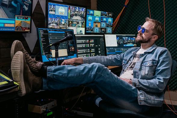 A photo of Cory Mcelrea, dressed as the wrestler Orange Cassidy in his trademark all-denim outfit & shared, sits behind a broadcast desk covered in screens with his feet up on the table.