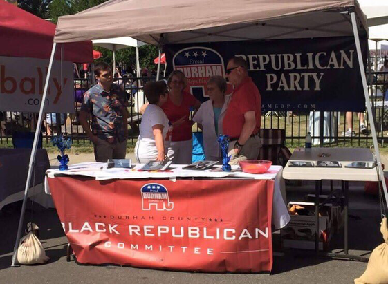Photo: An outdoor gathering (possibly a convention) where there’s an information table/tent (a “booth”) with a large table-length banner that reads, “DURHAM COUNTY BLACK REPUBLICAN COMMITTEE. There are five White people standing behind the table, but not a Black person ANYWHERE in the photo.
