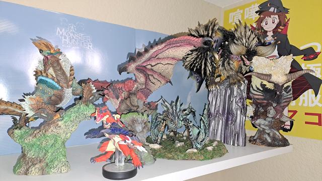 A series of statues on a shelf in front of a sky blue background and a print of fight club Mako from Kill la Kill. 
From left to right: Zinogre statue, Rathalos amiibo, Rathalos statue, Alduin sculpture, Nergigante statue, and Rajang statue.