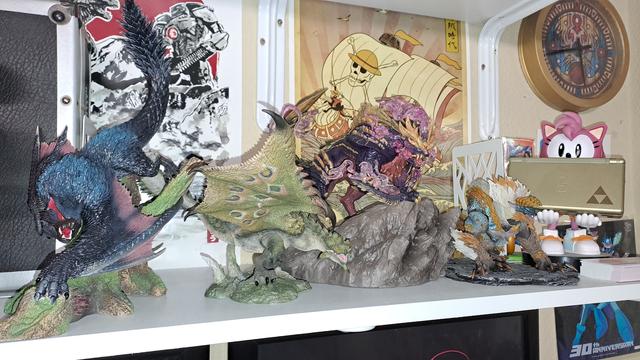 A series of statues on a shelf in front of a print of Godzilla vs Gypsy Danger, an ukiyoe style One Piece ship print, and a clock in the style of the clock tower from Majora's Mask.
From left to right: Nargacuga statue, Rathian statue, Magnamalo statue, Zinogre model, Classic Amy holding a Zelda styled DS lite.