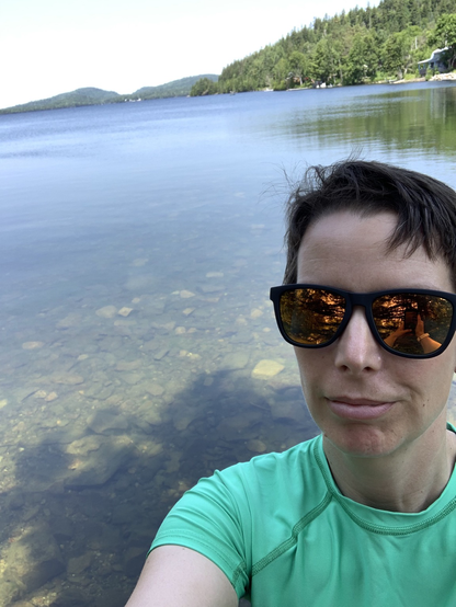 A short haired woman in reflective sunglasses, wearing a short sleeved rash guard, takes a selfie with a clear watered, calm lake stretching to the horizon behind her. The sun is shining down from a hazy blue sky.