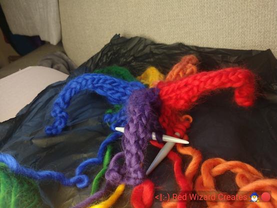 Picture of part of a knitted hat in six bright colours, the top of the hat forms a shape like some sort of sea creature with tentacles. Text at bottom reads: <|:-) Red Wizard Creates 🧙‍♂️🔴