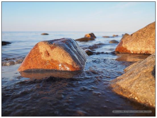 Eiszeitliche Findlinge liegen im flachen Wasser vor dem Ostseestrand, nur ganz kleine Wellen plätschern ans Ufer, die tiefstehende Abendsonne beleuchtet die Szene mit warm-rötlichem Licht; tiefe Kamerahaltung nur wenige Zentimeter über der Wasseroberfläche
