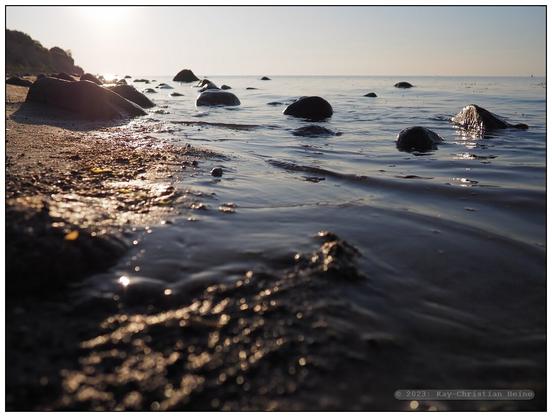Gegenlicht von der tiefstehenden Sonne, nur ganz kleine Wellen plätschern an den Strand, viele große Steine im flachen Wasser vor dem Spülsaum, tiefe Kamerahaltung nur wenige Zentimenter über der Wasseroberfläche - absolute Ruhe.