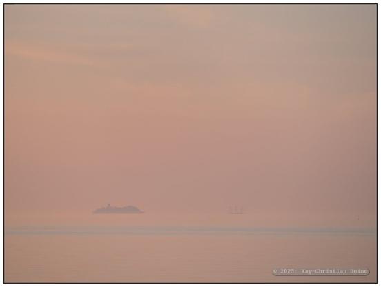 Von der tiefstehenden Sonne rosa gefärbter Dunst hebt die Grenze zwischen Ostsee, Horizont und Wasser auf, schemenhaft sind in der Ferne ein großes Fährschiff und ein Dreimastsegler auf dieser mit dem Teleobjektiv entstandenen Foto zu sehen.