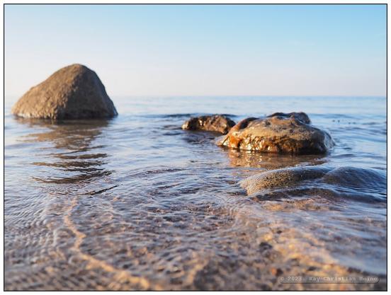 Eiszeitliche Findlinge liegen im flachen Wasser vor dem Ostseestrand, nur ganz kleine Wellen plätschern ans Ufer, die tiefstehende Abendsonne beleuchtet die Szene mit warm-rötlichem Licht; tiefe Kamerahaltung nur wenige Zentimeter über der Wasseroberfläche