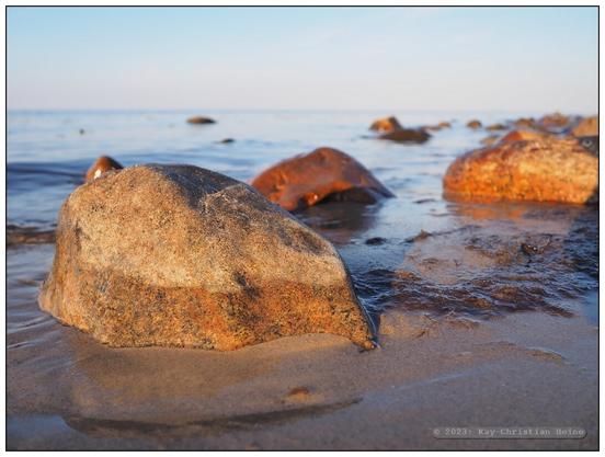 Eiszeitliche Findlinge liegen im flachen Wasser vor dem Ostseestrand, nur ganz kleine Wellen plätschern ans Ufer, die tiefstehende Abendsonne beleuchtet die Szene mit warm-rötlichem Licht; tiefe Kamerahaltung nur wenige Zentimeter über der Wasseroberfläche