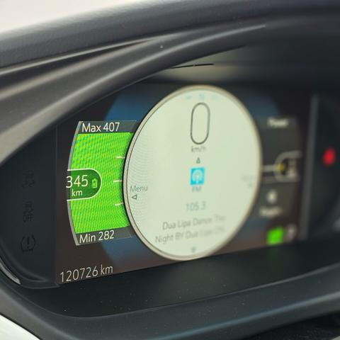 The binnacle of an electric vehicle with a full charge showing green charging bars and a white circle with current music and a red seatbelt sign