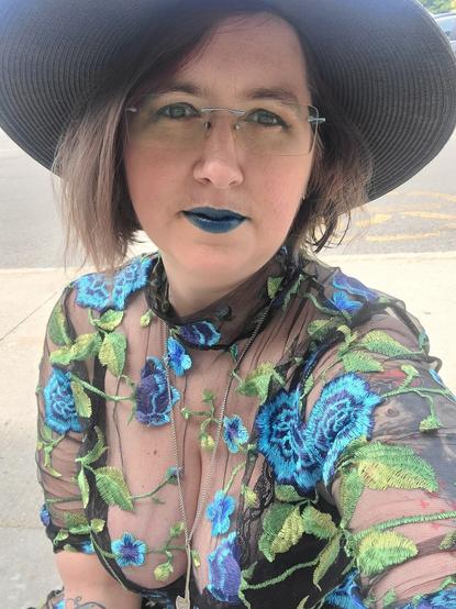 Wearing a semi-transparent black dress with blue flowers and green leaves broderie on it, wearing a black hat