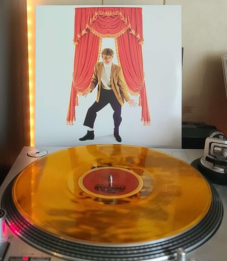 Image shows a turntable with a translucent orange vinyl record on the platter. Behind the turntable vinyl album outer sleeve is displayed. The front cover shows Modern nomad posing with his arms out in front of a current