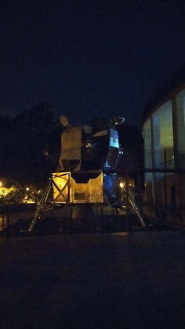 A model of a space craft (probably a planetary lander) standing on three feet outside a building in the dark.
