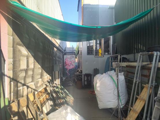 An alley behind a fence - photo taken while putting my arm through the bars - with two pieces. One: at the end of the alley, on some sort of construction a pink Friendly Hero, a cartoon figure, puffy looks, wearing a pink cape, yellow closed hood, white face, looking startled/amazed, red heart on its chest, right hand up in the air, left arm around the neck of the pink flamingo that its sitting on. Three exclamation marks above its head, text balloon to the right of its head, with a red heart, and three flying birds in it.
Two: on the left wall of the alley a piece that spells "Utrecht" in capital letters, in 3D as if the letters were cut out in the wall. The letters themselves also have a 3D effect, seemingly protruding further or less from the wall. This second piece is likely of JanIsDeMan, very alike to the piece on the picture linked to in the post, and having his office/atelier close by.
In the alley there is a collection of building materials, partly in front of the pieces.