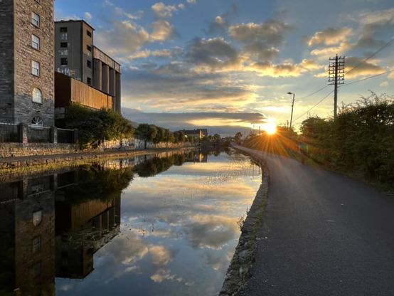 Sunsetting against a blue sky with some clouds. The sky and the tall buildings are mirrored on a perfectly still canal.