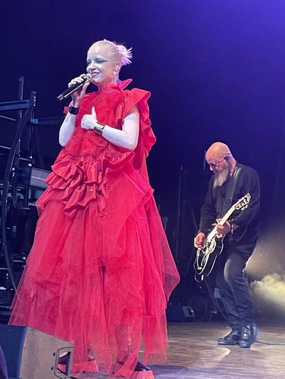 Shirley Manson and Steve Marker of Garbage during one of their recent shows. Shirley is in a frilly red dress with her hair pulled back into a short ponytail, singing into a handheld mic. Steve Marker wears all black and plays a black guitar with white edges in the background behind her. The stage lighting is white light with a little purple in the upper right.