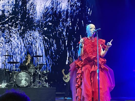 Shirley Manson sings into the mic that is on her mic stand. She wears a very frilly red dress. Touring bassist Daniel Schulman and drummer Butch Big are in the background behind her. Butch is about to hit a cymbal. There is a projected abstract image behind them. It has a black background with white, blue, and purpley blue lines streaked across like a painting.