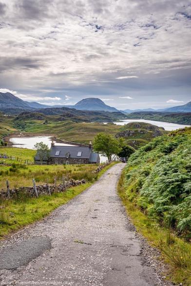 A photo of the Scottish highlands with a road leading to a cottage half way down the hill. There’s lush green foliage on with side of the road with stonewall and fencing on one side and green fern on the other. The road leads to a winding Loch and sandy shoreline past the cottage with high hills in the foreground and cloudy but bright skyline.